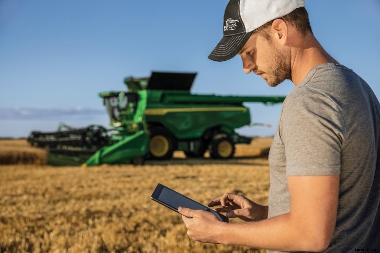 A man stands in a field with an iPad with a green John Deere combine behind him